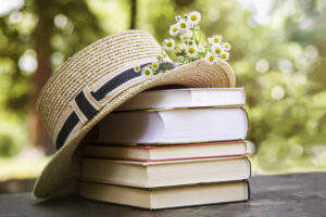 A stack of books on an old wooden table next to a straw hat of a canoe and a bouquet of wild flowers.